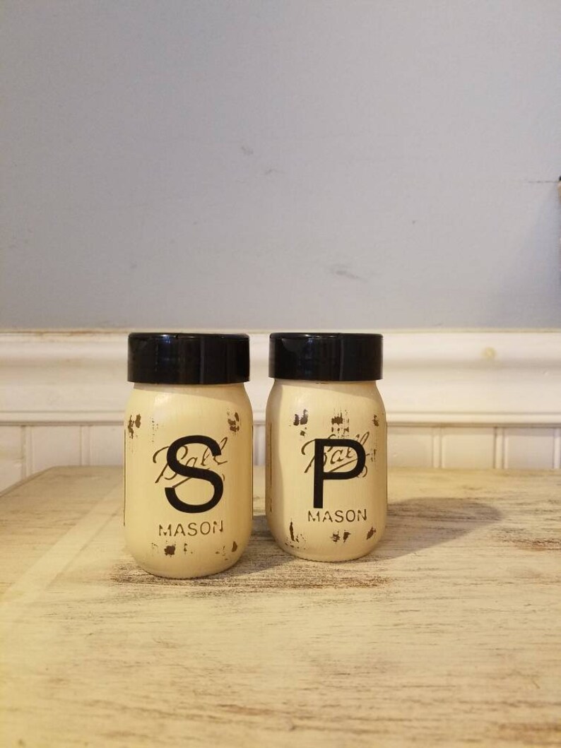 May include: Two vintage-style glass salt and pepper shakers with black lids. The shakers are labeled "S" and "P" with the word "MASON" underneath. The shakers are sitting on a light wood surface.