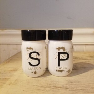May include: White painted mason jars with black lids and gold lettering. The jars are labeled "S" and "P" for salt and pepper.