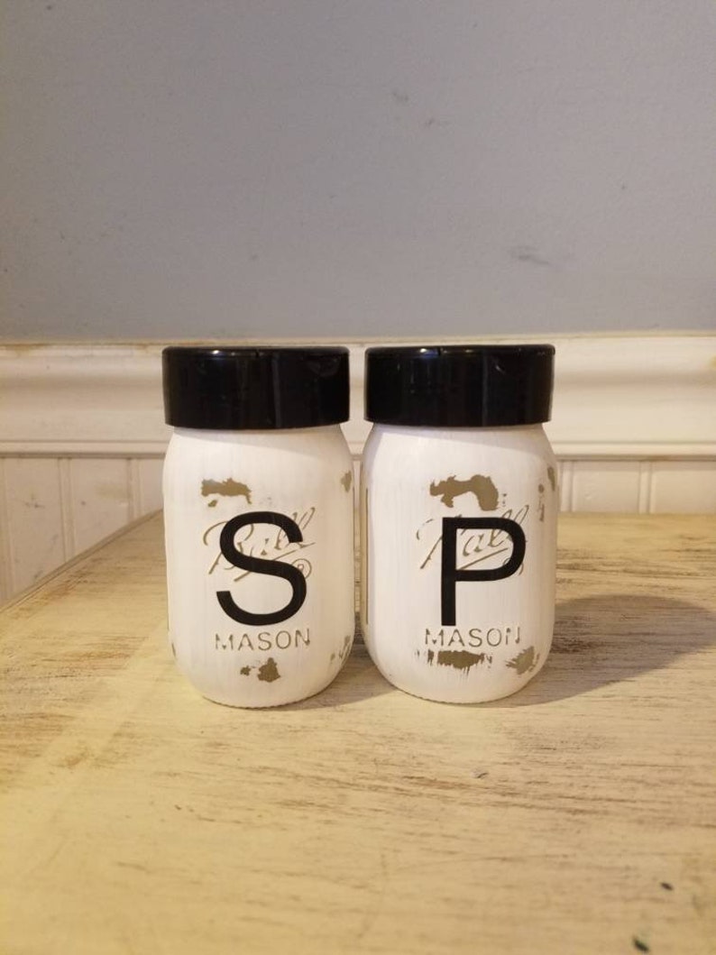 May include: White painted mason jars with black lids and gold lettering. The jars are labeled "S" and "P" for salt and pepper.