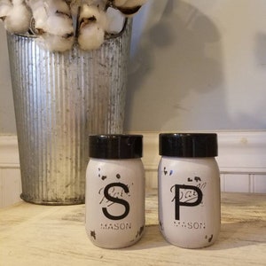 May include: White painted mason jars with black lids and black lettering that reads "S" and "P" for salt and pepper. The jars are sitting on a light wood surface.