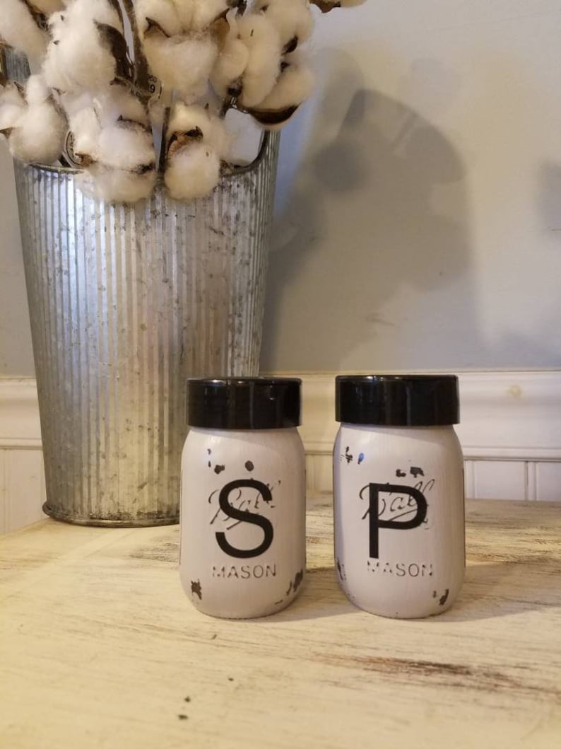 May include: White painted mason jars with black lids and black lettering that reads "S" and "P" for salt and pepper. The jars are sitting on a light wood surface.