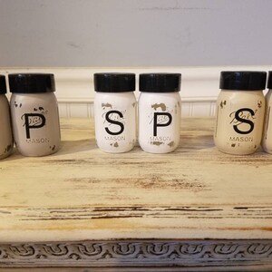 May include: Six small glass jars with black lids and white labels. The labels have the letters "S", "P", and "S" written in black. The jars are arranged in a row on a light brown wooden surface.
