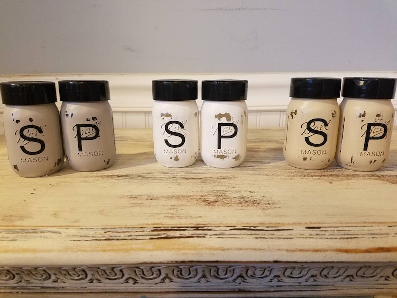 May include: Six small glass jars with black lids and white labels. The labels have the letters "S", "P", and "S" written in black. The jars are arranged in a row on a light brown wooden surface.