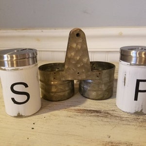 K&ouml;nnte beinhalten: Wei&szlig;e Salz- und Pfefferstreuer mit schwarzer Beschriftung "S" und "P". Die Streuer befinden sich in einem Metallhalter mit Griff.