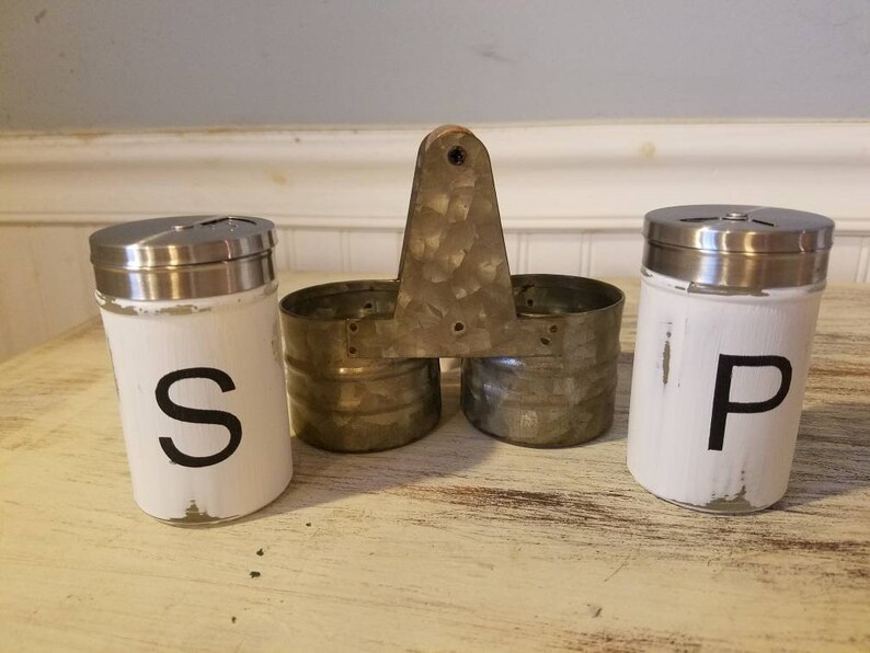 K&ouml;nnte beinhalten: Wei&szlig;e Salz- und Pfefferstreuer mit schwarzer Beschriftung "S" und "P". Die Streuer befinden sich in einem Metallhalter mit Griff.
