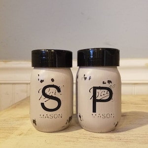 May include: Two white mason jars with black lids and black lettering. The jars are labeled "S" and "P" for salt and pepper.