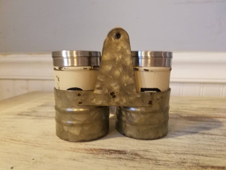 K&ouml;nnte beinhalten: Ein Set aus zwei Salz- und Pfefferstreuern aus Metall mit einer patinierten, verzinkten Oberfl&auml;che. Die Streuer werden von einem Metallgestell mit Griff zusammengehalten.