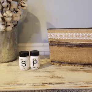 May include: Two small white glass salt and pepper shakers with black lids and labels that say "S" and "P" with the word "Mason" underneath. The shakers are on a white wooden surface with a burlap and lace basket behind them.