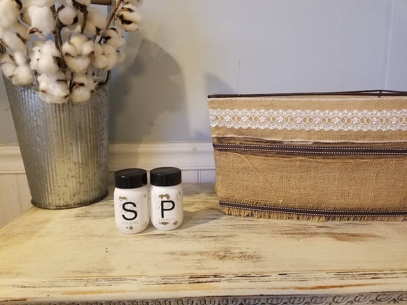 May include: Two small white glass salt and pepper shakers with black lids and labels that say "S" and "P" with the word "Mason" underneath. The shakers are on a white wooden surface with a burlap and lace basket behind them.