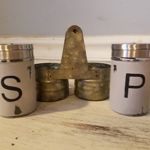 K&ouml;nnte beinhalten: Ein Set aus zwei Salz- und Pfefferstreuern aus Metall mit einer abgenutzten wei&szlig;en Oberfl&auml;che. Die Streuer sind mit "S" und "P" beschriftet und an einem Metallst&auml;nder mit einem Holzgriff befestigt.