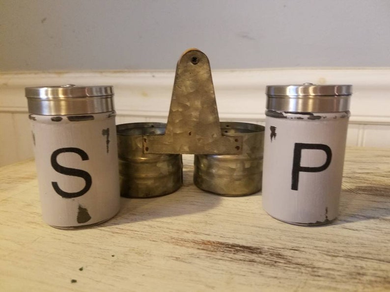 K&ouml;nnte beinhalten: Ein Set aus zwei Salz- und Pfefferstreuern aus Metall mit einer abgenutzten wei&szlig;en Oberfl&auml;che. Die Streuer sind mit "S" und "P" beschriftet und an einem Metallst&auml;nder mit einem Holzgriff befestigt.