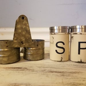 K&ouml;nnte beinhalten: Ein Set aus zwei wei&szlig;en Salz- und Pfefferstreuern mit schwarzer Beschriftung "S" und "P" auf einer rustikalen Holzoberfl&auml;che. Die Streuer befinden sich in einem Metallhalter mit Griff.