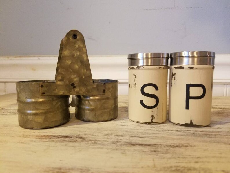 K&ouml;nnte beinhalten: Ein Set aus zwei wei&szlig;en Salz- und Pfefferstreuern mit schwarzer Beschriftung "S" und "P" auf einer rustikalen Holzoberfl&auml;che. Die Streuer befinden sich in einem Metallhalter mit Griff.