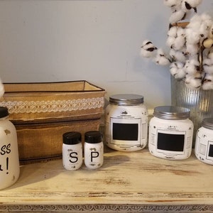 May include: White painted jars with black chalkboard labels, used for storing spices or other kitchen items. The jars are arranged on a white shelf with a brown burlap box and a white tissue box holder that says "Bless You!"