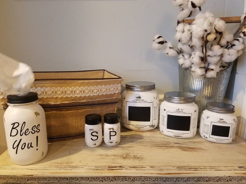May include: White painted jars with black chalkboard labels, used for storing spices or other kitchen items. The jars are arranged on a white shelf with a brown burlap box and a white tissue box holder that says "Bless You!"