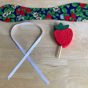 May include: A blue fabric headband with a strawberry and floral pattern, a white ribbon, and a red wooden strawberry on a stick. The strawberry has a green top and is painted with small white dots.