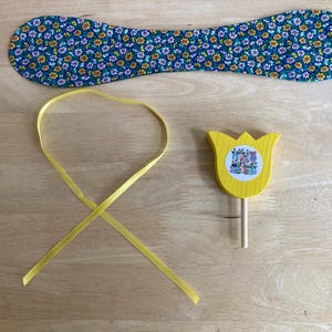 May include: A blue floral fabric item, a yellow ribbon, and a yellow wooden tulip with a circular sticker are arranged on a wooden surface. The fabric item is a long, curved shape. The tulip has a wooden stem.