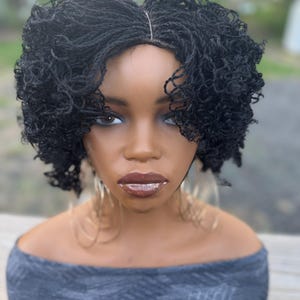 May include: A black micro-braided wig styled with curly ends, displayed on a mannequin head. The mannequin has dark skin, full lips, and large hoop earrings. The wig is a short, textured style.