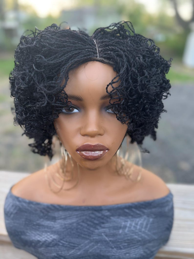 May include: A black micro-braided wig styled with curly ends, displayed on a mannequin head. The mannequin has dark skin, full lips, and large hoop earrings. The wig is a short, textured style.