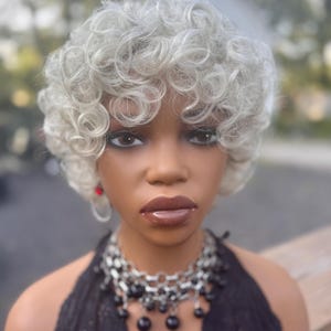May include: A mannequin head with a short, curly, silver wig. The mannequin wears a black halter top, a silver and black beaded necklace, and red earrings. The mannequin has dark eye makeup and brown lipstick.
