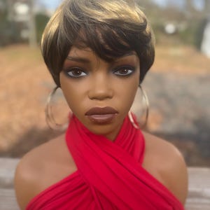 May include: A mannequin head with a short, layered wig in shades of brown and blonde. The mannequin has dark eye makeup, full lips, and large gold hoop earrings. A bright red, draped fabric covers the mannequin's chest.