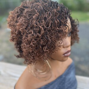 May include: A short, curly wig with a blend of brown and black tones. The wig is styled with tight, defined curls and is worn on a person with a gray off-the-shoulder top. Large gold hoop earrings are visible.
