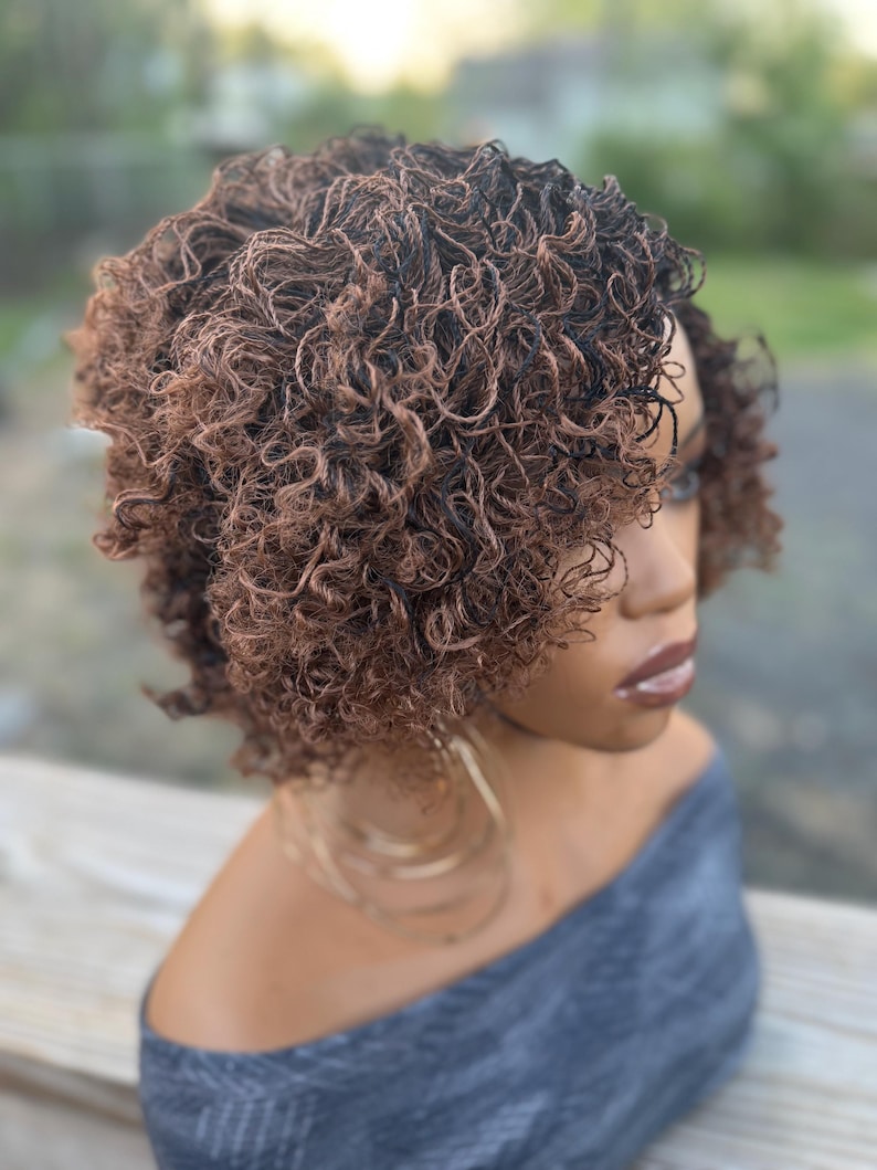 May include: A short, curly wig with a blend of brown and black tones. The wig is styled with tight, defined curls and is worn on a person with a gray off-the-shoulder top. Large gold hoop earrings are visible.