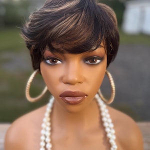 May include: A mannequin head with a short, dark brown wig with blonde highlights. The mannequin wears gold hoop earrings and a pearl necklace. The mannequin has brown eyes and glossy brown lipstick.