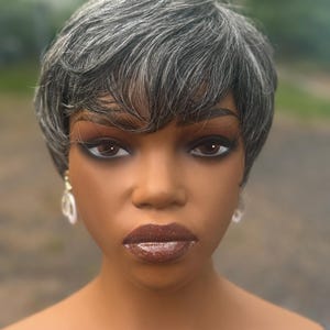 May include: A mannequin head with a short, gray wig styled with soft waves. The mannequin has dark brown eyes, defined eyebrows, and shimmering brown lipstick. Clear, circular earrings dangle from the ears.