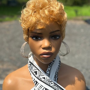 Op de afbeelding: Een kort, krullend, blond pruik met een scheiding aan de zijkant. De pruik zit op een mannequinhoofd met een wit en zwart gepatroneerde sjaal.