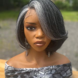 Könnte beinhalten: Eine kurze, gestufte Bob-Perücke mit einer Mischung aus dunkelanthrazitfarbenem und silbergrauem Haar. Die Perücke hat einen seitlich gescheitelten Pony und einen Lace-Front-Ansatz. Die Schaufensterpuppe trägt einen schwarz-grau gemusterten Schal.
