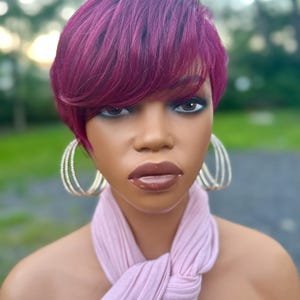 May include: A mannequin head with a short, layered, burgundy wig. The mannequin wears large, gold hoop earrings and a light pink scarf-like top. The background is blurred with green and gray tones.