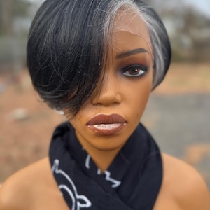 Puede incluir: Una cabeza de maniquí con una peluca corta negra con una raya a un lado y reflejos grises. El maniquí tiene maquillaje completo, incluyendo sombra de ojos oscura y lápiz labial marrón. Una bufanda negra con un patrón blanco está drapeada alrededor del cuello.