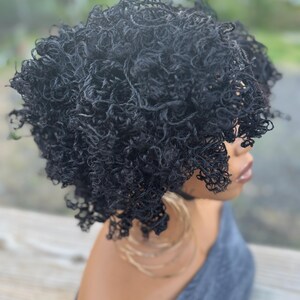 May include: A black curly wig with a natural-looking texture. The wig is styled in a short, voluminous afro. The person is wearing large gold hoop earrings and a gray top. The background is blurred, suggesting an outdoor setting.