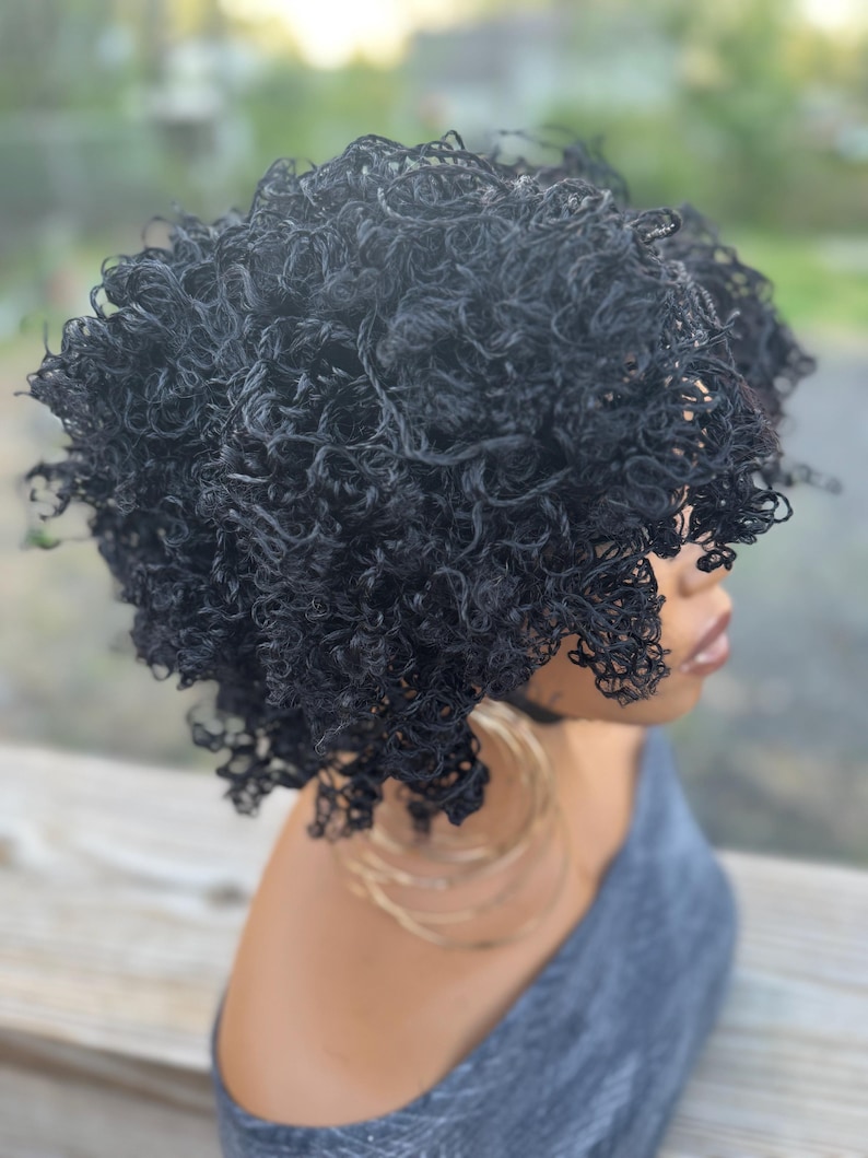 May include: A black curly wig with a natural-looking texture. The wig is styled in a short, voluminous afro. The person is wearing large gold hoop earrings and a gray top. The background is blurred, suggesting an outdoor setting.