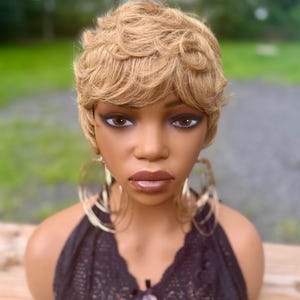 May include: A mannequin head with a short, wavy, light brown wig. The mannequin wears large gold hoop earrings and a black lace top with a pink and brown leopard print. The background is blurred, with green grass and a gray road.