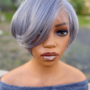 Op de afbeelding: Een korte, gelaagde zilvergrijze pruik met een zijwaartse pony en een zichtbare lace front haarlijn. De pruik is gestyled op een etalagepophoofd met donkerbruine ogen en glanzende, glitterende bruine lippen, gekleed in een blauwe top.