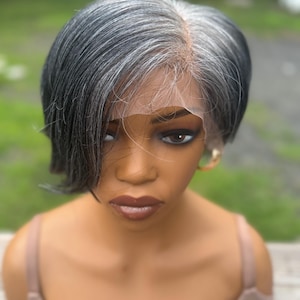 May include: A short, layered wig featuring a blend of black and silver-gray hair with a side part. The wig is displayed on a mannequin head and includes a visible sheer lace front hairline that extends across the forehead for a natural appearance.