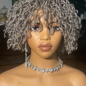 Puede incluir: Una peluca gris plateada con cabello corto, rizado y texturizado. La peluca está peinada con raya lateral y flequillo.