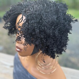 May include: A black curly wig with a short, layered style. The wig features a combination of tight curls and braided sections, creating a textured look. The wig is styled on a mannequin head, with gold hoop earrings visible.