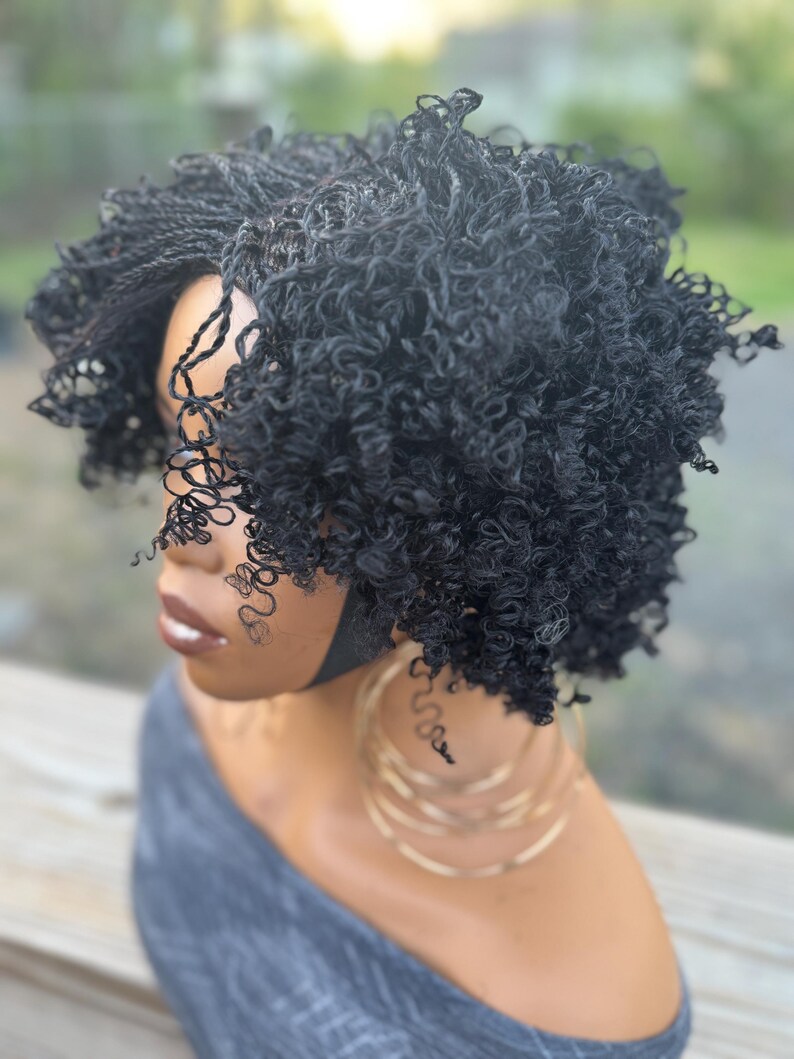 May include: A black curly wig with a short, layered style. The wig features a combination of tight curls and braided sections, creating a textured look. The wig is styled on a mannequin head, with gold hoop earrings visible.