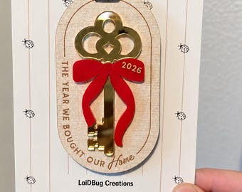 Puede incluir: Un adorno ovalado de madera con una llave de espejo dorada, un lazo rojo y el texto 2026. El adorno está sujeto a una tarjeta blanca con iconos de mariquitas y el texto Holiday Collection, Handmade With Love, LaidBug Creations.