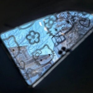 May include: A blue car window with a Hello Kitty sticker design. The sticker features a white Hello Kitty outline with black details and a heart.