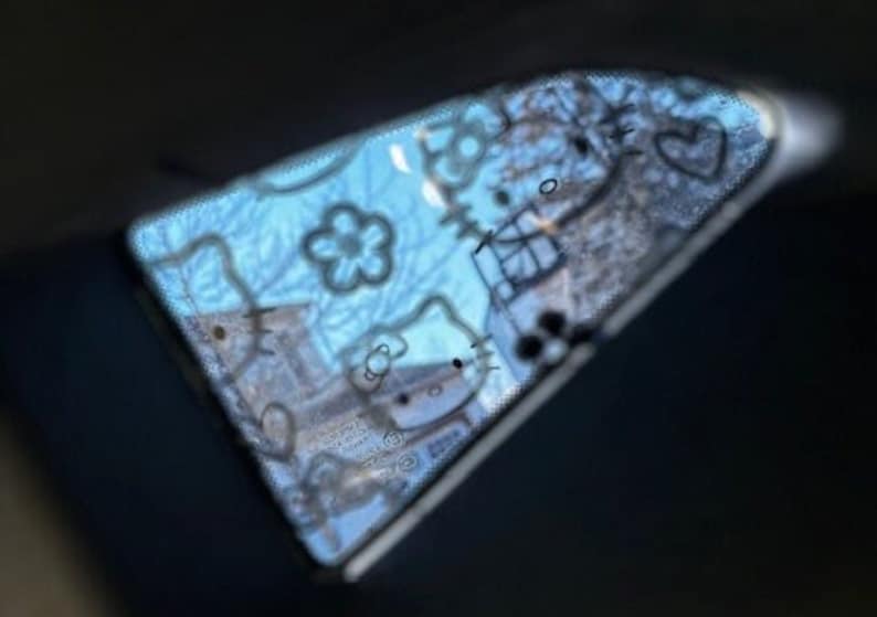 May include: A blue car window with a Hello Kitty sticker design. The sticker features a white Hello Kitty outline with black details and a heart.
