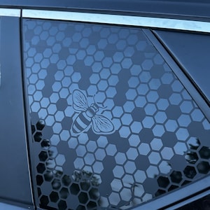 May include: A black car window with a honeycomb pattern and a bee graphic. The bee is in the center of the window and is facing to the right. The honeycomb pattern is in the background and is made up of small hexagons.