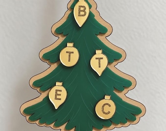 Puede incluir: Un adorno con forma de árbol de Navidad hecho de madera y acrílico verde, con cinco bolas doradas con las letras B, T, T, E y C. Una base roja en la parte inferior muestra el texto blanco 2026. Una cinta blanca está unida a la parte superior.