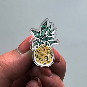 May include: A yellow and green pineapple pin with a clear acrylic backing.