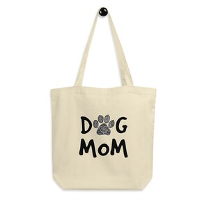 K&ouml;nnte beinhalten: Eine beige Canvas-Tragetasche mit schwarzem Text, der "DOG MOM" lautet, und einem schwarzen Pfotenabdruck mit einem detaillierten Design in der Mitte des "O".