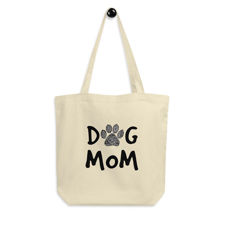 K&ouml;nnte beinhalten: Eine beige Canvas-Tragetasche mit schwarzem Text, der "DOG MOM" lautet, und einem schwarzen Pfotenabdruck mit einem detaillierten Design in der Mitte des "O".