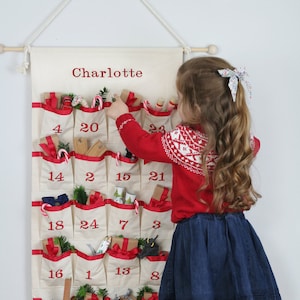 Fabric advent calendar with gusseted pockets trimmed with red ribbon. The calendar hangs from a cotton cord on a wooden rod. 
The numbers are placed randomly and embroidered in red thread.
The calendar can be personalised.